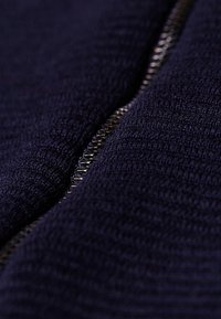 Närbild av en delvis zippad metalldragkedja på mörkt texturerat tyg, som framhäver dragkedjans tänder och det stickade materialet.