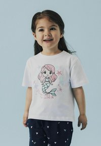 Camiseta blanca de algodón con un gráfico colorido de sirena con cabello rosa, rodeada de estrellas y elementos marinos, combinada con pantalones oscuros estampados.