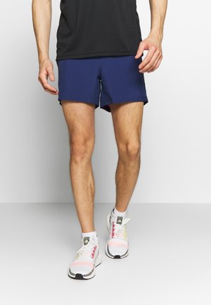 Personne portant un short de sport bleu marine, un t-shirt noir, des chaussettes blanches et des chaussures de course blanches avec des accents roses et verts, debout sur un sol gris.