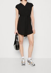 Svart kort klänning med v-ringning, korta ärmar och knytliv. Kombinerad med en glänsande svart väska och grå sneakers med orange detaljer.