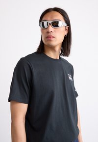 Zonnebril met een glanzend witte brilrand en spiegelende lenzen, gedragen met een fitted zwarte katoenen t-shirt. Het haar is lang en donker.
