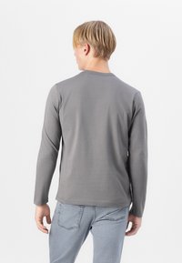 Cămașă gri cu mâneci lungi, confecționată dintr-un material moale, având guler rotund și un design simplu. Poartă blugi albaștri deschiși, prezentată din spate.