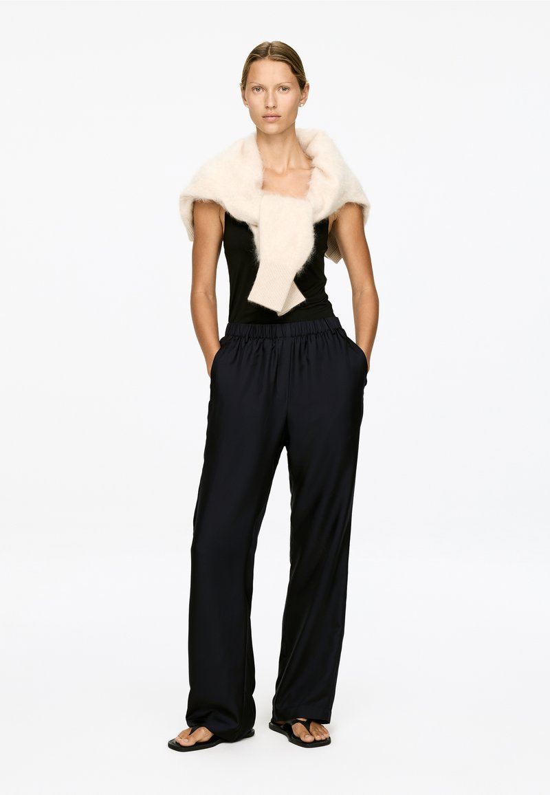 Femme portant un haut sans manches noir, un pantalon noir ample, des sandales et un pull en peluche crème drapé sur les épaules, se tenant avec les mains dans les poches.