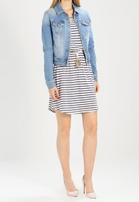 Vaaleansininen farkkutakki nappisuljennuksella, jossa on kaksi rintataskua, yhdistettynä raidalliseen hihattomaan mekkoon ja vaaleanpuniin korkokenkiin.