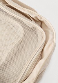 Μπεζ τσάντα αποθήκευσης από πλέγμα με επένδυση από καπιτονέ ύφασμα, με φερμουάρ και λεία υφή. Εσωτερική τσέπη ορατή μέσα από το πλέγμα.