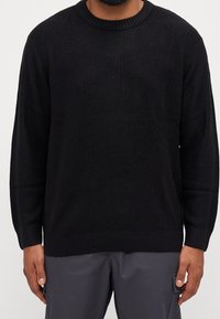 Pull noir en tricot avec un col rond, des poignets côtelés et un motif texturé. Coupe décontractée, matériau léger.
