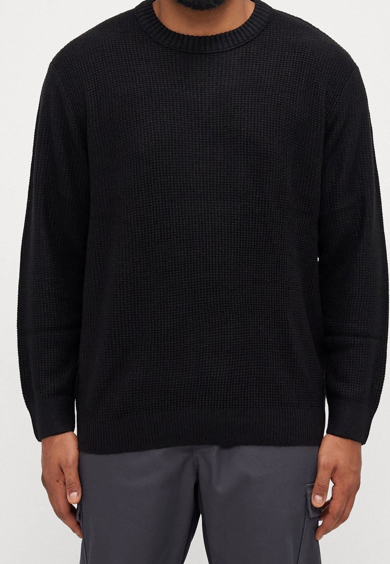 Pull noir en tricot avec un col rond, des poignets côtelés et un motif texturé. Coupe décontractée, matériau léger.