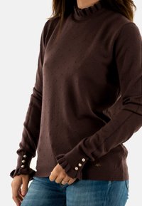 Maglione marrone con motivo a pallini testurizzati, colletto arricciato e polsini con dettagli in perle. Indossato con jeans blu; materiale in maglia morbida.
