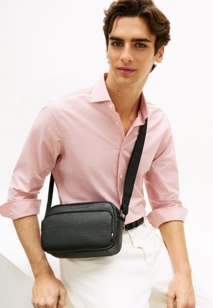 Giovane uomo che indossa una camicia rosa chiaro e pantaloni bianchi, con una borsa a tracolla nera con pannello frontale texturizzato.