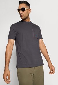 Tricou gri cu buzunar și detalii logo verde-albastru, mâneci scurte, textură moale. Purtați cu ochelari de soare bej și pantaloni verzi măslinii.