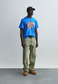 Μπλε βαμβακερό μπλουζάκι με ροζ γραφικό κείμενο, συνδυασμένο με πράσινο ριγέ παντελόνι και καφέ μπότες. Το μοντέλο φοράει σκούρο καπέλο.