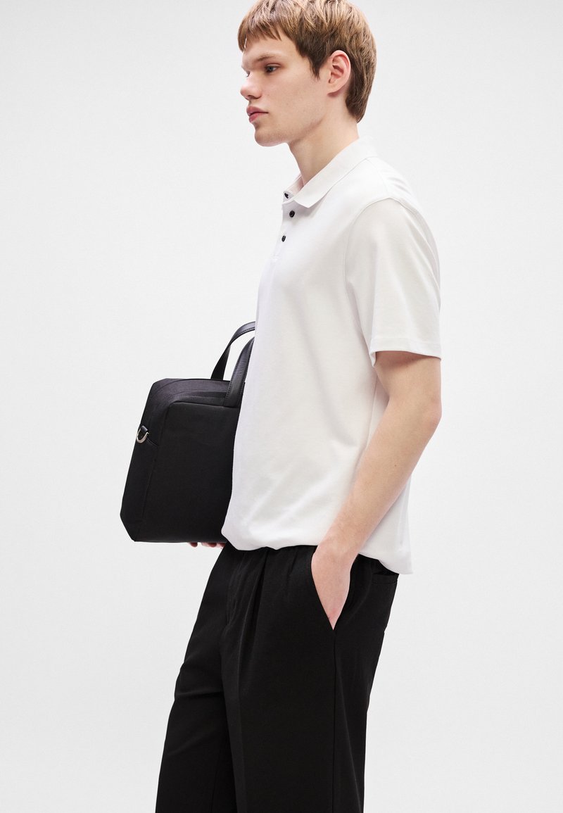 Chemise polo blanche avec deux boutons noirs, associée à un pantalon noir ample. La personne tient un tote bag noir avec de courtes poignées.