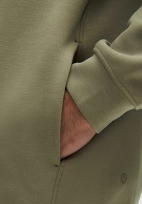 Sweatshirt vert olive avec une texture lisse, présentant une poche latérale et des poignets ajustés. Un petit logo est visible près de l'ourlet.