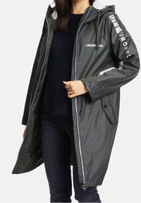 Dlouhá, voděodolná pláštěnka tmavě zelené barvy s bílým logem. Má stahovací kapuci, zip na přední straně a kapsy. Hladká struktura.