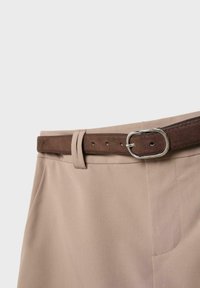 Beżowe spodnie z bocznymi kieszeniami, zapinane brązowym zamszowym paskiem z srebrną owalną klamrą, na jednolitym jasnym tle.