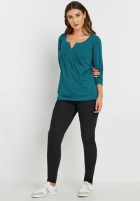 Maglietta teal a maniche 3/4 con scollatura rotonda, dettagli con bottoni e pieghe a pizzico, abbinata a jeans skinny neri e scarpe da ginnastica bianche.