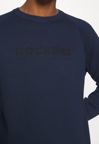 Námořnická modrá mikina se žebrovaným kulatým výstřihem a raglánovými rukávy. Na přední straně se nachází černě tištěné logo "DOCKERS ESTABLISHED 1986".