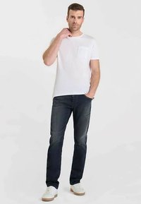 T-shirt blanc à col rond avec une poche poitrine, associé à un jean foncé. Le modèle porte des baskets blanches ; la tenue est décontractée et confortable.