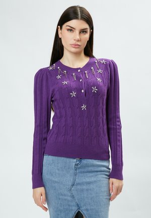 Lila gestrickter Pullover mit Kabelmuster, verfügt über dekorative silberfarbene Akzente und Knöpfe sowie gerippte Bündchen. Getragen mit einem Jeansrock.