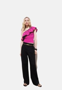 Fuchsia Oberteil mit einem Schulterträger und Rüschendetail; kombiniert mit schwarzen weiten Hosen und einem goldenen Kettengürtel. Sandalen vervollständigen das Outfit.