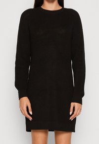 Abito nero lavorato a maglia con maniche lunghe, scollatura rotonda, polsini e orlo a costine. Ha una texture spessa e una vestibilità ampia.