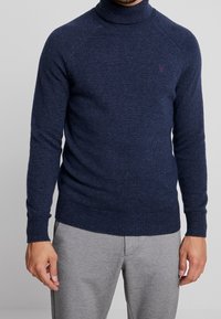 Pull à col roulé bleu marine foncé avec texture côtelée, manches longues et coupe ajustée. Présente un petit emblème sur la poitrine. Associé à un pantalon gris.