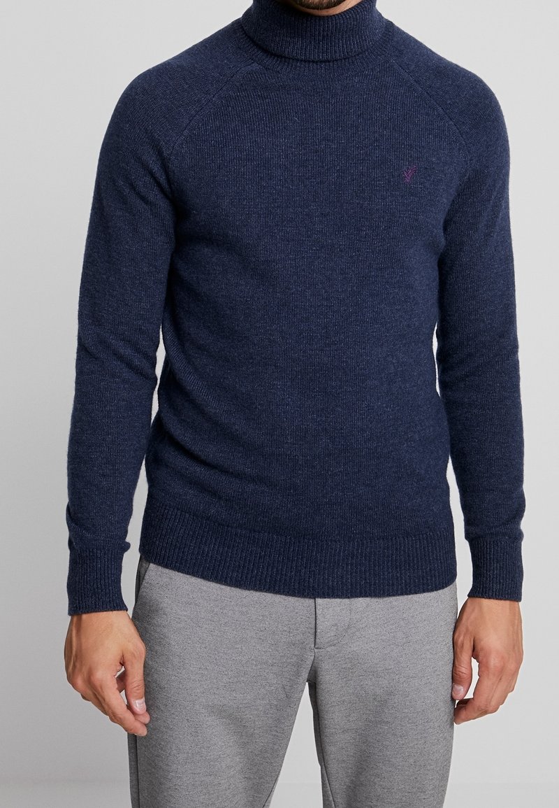 Pull à col roulé bleu marine foncé avec texture côtelée, manches longues et coupe ajustée. Présente un petit emblème sur la poitrine. Associé à un pantalon gris.