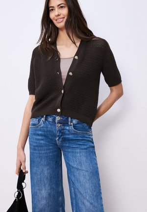 Femme souriante portant un cardigan court en maille marron foncé sur un haut beige et un jean taille haute bleu, tenant un sac à main noir.