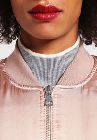Rosa satin bomberjacka med en silverfärgad dragkedja, med en grå ribbad polokrage under. Slät textur, kontrasterande krage.