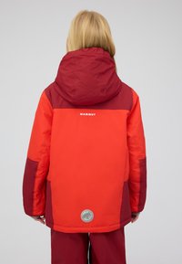Veste rouge vif avec des accents rouge foncé, capuche et logo au dos. Tissu durable avec une texture lisse et un emblème circulaire à l'ourlet.