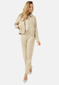 Giacca bomber beige con zip frontale, polsini elasticizzati, pantaloni coordinati e décolleté a punta con fiocchi decorativi. Tessuto morbido.