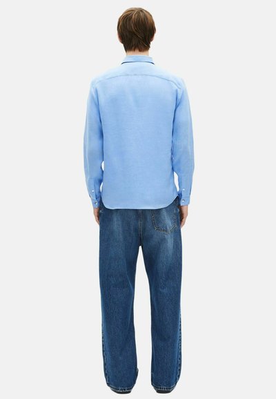 Άτομο γυρισμένο προς τα πίσω που φοράει γαλάζιο πουκάμισο με μακριά μανίκια και φαρδιά σκούρα μπλε τζιν, μπροστά σε απλό λευκό φόντο.