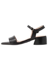 Mustan nahkakengän tasapohjainen, avonainen kärki, säädettävä nilkkahihna ja pieni, paksu korko. Sisältää kuusikulmaisen soljen korostuksen.