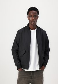 Svart bomberjacka med ribbad krage och muddar, framtagningsdragkedja och två sidofickor, bärs över en vit t-shirt och grå jeans.