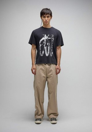 Jeune homme debout portant un t-shirt graphique noir avec le texte "Torment", un pantalon beige à jambes larges et des baskets noires sur un fond gris uni.