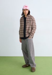 Camisa de franela marrón a cuadros sobre una camiseta negra, pantalones de pana grises y zapatos marrones. Gorro rosa con detalle en texto. Fondo de suelo verde.
