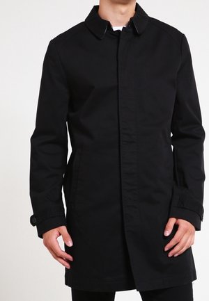 Man wearing a black long coat with a collar, buttoned cuffs, and side pockets, standing against a plain light background.