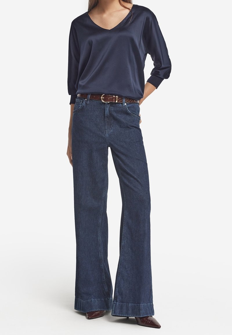Femme portant une blouse en satin bleu marine à manches longues, un jean en denim foncé taille haute à jambes larges, une ceinture marron et des chaussures à bout pointu.