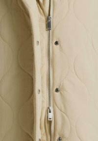 Beiges gesteppter Stoff mit Metallreißverschluss und Druckknöpfen entlang der mittleren Öffnung eines Jacketts oder Westenbekleidung.