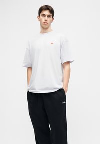 Νεαρός με κοντά μαλλιά στέκεται με τα χέρια στις τσέπες, φορώντας λευκό μπλουζάκι και μαύρη φόρμα με μικρά λογότυπα μάρκας.