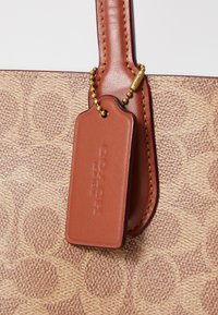 Tan fabric tote bag with a signature pattern, featuring a brown leather tag attached by a gold chain, accented with contrasting stitching.