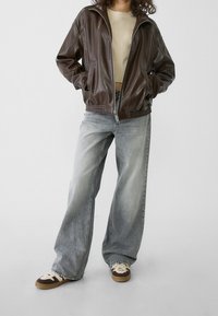 Chaqueta marrón de cuero sintético con cremallera y bolsillos laterales, combinada con vaqueros anchos de color gris claro y zapatillas marrones con detalles en crema.