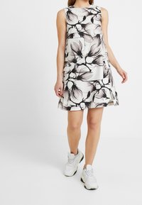 Žena v bezramínkových bílých šatech s černými květinovými vzory a bílých chunky teniskách, stojící na pozadí z čistě bílé barvy.