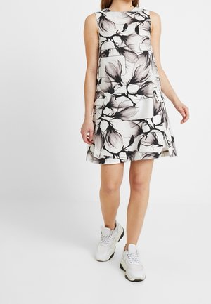 Kvinna som bär en ärmlös vit klänning med svarta blommönster och vita chunky sneakers, står mot en enkel vit bakgrund.