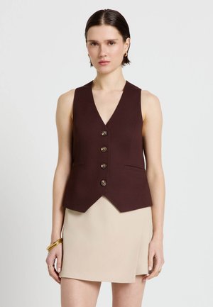Giovane donna che indossa un gilet senza maniche marrone scuro con bottoni e una mini gonna beige, accessoriata con braccialetti e anelli d’oro.