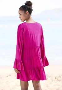 Vestido de fucsia a la altura de la rodilla con un corte relajado, que presenta mangas de campana y detalles de volantes en capas. La tela suave complementa los entornos de playa.