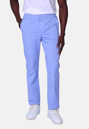 Homme portant un pantalon bleu clair à coupe droite, des baskets blanches et une chemise blanche rentrée, se tenant debout avec une main dans la poche.