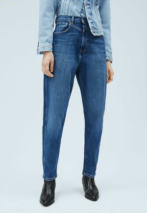 Persoon die een high-waisted blauwe spijkerbroek, een licht denim jack en zwarte puntige enkellaarsjes draagt, staand tegen een eenvoudige lichte achtergrond.