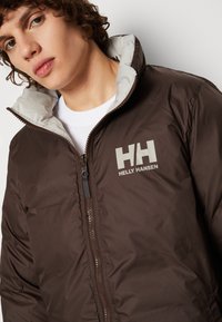 Hnědá izolační bunda se šedou vnitřní podšívkou, z přední strany na zip a s logem "HH" na prsou. Hladká, voděodolná tkanina.