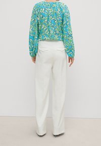 Blouse à manches longues, avec un motif bleu et vert, dotée d'un col froncé, associée à un pantalon taille haute, blanc et ajusté, avec des poches arrière.